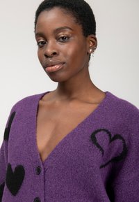 Primo piano di una donna sicura di sé che indossa un cardigan viola con motivi a cuore neri e orecchini di perle, su uno sfondo neutro.