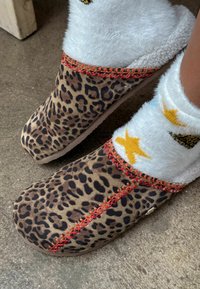 Leopardmønstrede træsko med et blødt, pelslignende indre, med røde sting langs åbningen, kombineret med hvide sokker, der viser farverige mønstre.