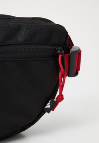 Sort stofpose med en tekstureret overflade. Har røde snore detaljer, klipslukning og et lille mærke med et logo på siden.