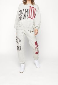 Felpa grigia chiara e pantaloni abbinati, con testo audace in nero e rosso. Design casual con una vestibilità rilassata e polsini elasticizzati.