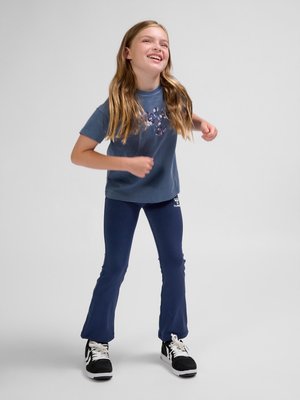 Młoda dziewczyna uśmiechająca się i poruszająca się swobodnie, ubrana w niebieski t-shirt, granatowe spodnie i czarno-białe trampki na jednolitym tle.