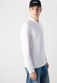 Langermet hvit polo skjorte med krage, brukt med blå denim bukser. Capsen har "HUGO" brodert i hvitt. Enkelt design, rene linjer.
