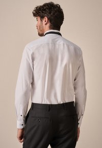 Bílá košile s dlouhými rukávy, s černým límcem a kontrastními černými manžetami, v kombinaci s tmavými kalhotami. Čistý, střihaný střih.