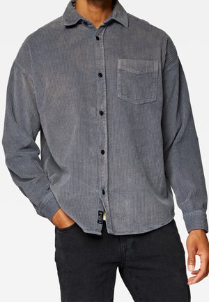 Mand iført en grå fløjls-skjorte med brystlomme og sort jeans, hånden i lommen, mod en ensfarvet baggrund.