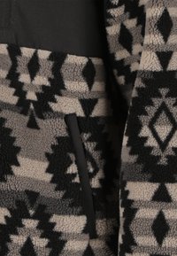 Fleecejacke in Schwarz, Beige und Grau mit geometrischen Mustern. Verfügt über eine Stofftasche und eine weiche, strukturierte Oberfläche.