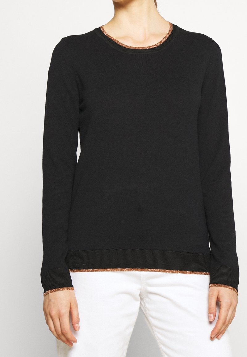 Femme portant un pull noir ajusté à manches longues avec une bordure cuivre au col, aux poignets et à l'ourlet, associé à un pantalon blanc.