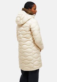 Beige gewatteerde pufferjas met capuchon, met een golfpatroon en een zachte textuur. Het model draagt een donker laagje eronder.