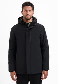 Man in een zwarte winterjack met een capuchon, rits en drukknopen, staande tegen een effen witte achtergrond.