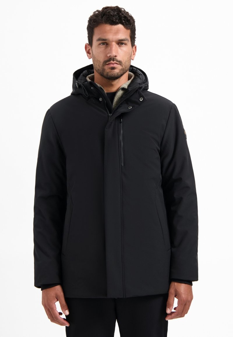 Man in een zwarte winterjack met een capuchon, rits en drukknopen, staande tegen een effen witte achtergrond.