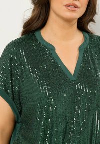 Top verde a maniche corte con scollatura a V, decorato con paillettes per un effetto texture. Tessuto liscio con un effetto scintillante.