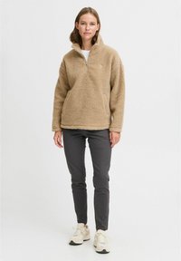 Pull en polaire beige avec col zippé à moitié et surface texturée, associé à un pantalon sombre et des baskets claires. Style décontracté.