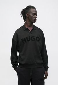 Férfi rasztahajjal, fekete HUGO pulóvert és hozzá illő nadrágot visel, zsebében tartja a kezét, oldalra néz.