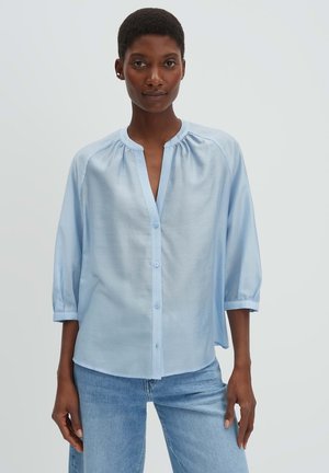 Femme aux cheveux courts portant un chemisier bleu clair à boutons et un jean bleu, debout devant un fond blanc uni.