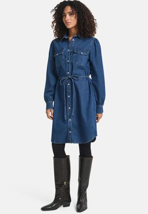 Denim shirt dress with long sleeves, button-up front, two chest pockets, cinched waistline, and a knee-length cut. Worn with black knee-high boots.