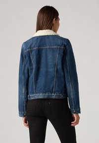 Denimjakke, mørkeblå, med en hvit shearlingkrage, knappede mansjetter og synlige sømdetaljer på baksiden og skuldrene.