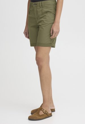 Egy személy, aki olívazöld térdig érő rövidnadrágot és barna színű, csatos részlettel ellátott nyersbőr bebújós cipőt visel, hófehér háttér előtt áll.