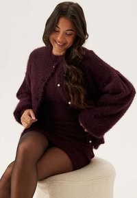 Burgundy fluffig cardigan med vida ärmar över en matchande figurnära klänning. Har pärlaccents på cardiganen. Mjuk textur och varm design.