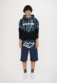 Black hoodie with blue lightning pattern and white text, paired with blue denim shorts featuring a white cartoon graphic. White sneakers, gray socks.