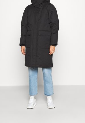 Personne portant un long manteau noir matelassé avec des poches à rabat, un jean bleu clair raccourci et des baskets blanches, se tenant devant un fond uni.