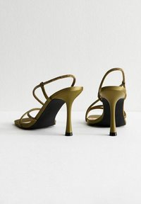 Sandali con tacco alto color verde oliva con sottili cinturini, decorati con accenti metallici, caratterizzati da una silhouette elegante e una suola nera texturizzata.