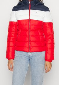 Person i en pufferjacka med marinblå, vit och röd horisontell randning och ljusblå jeans, stående mot en enfärgad bakgrund.