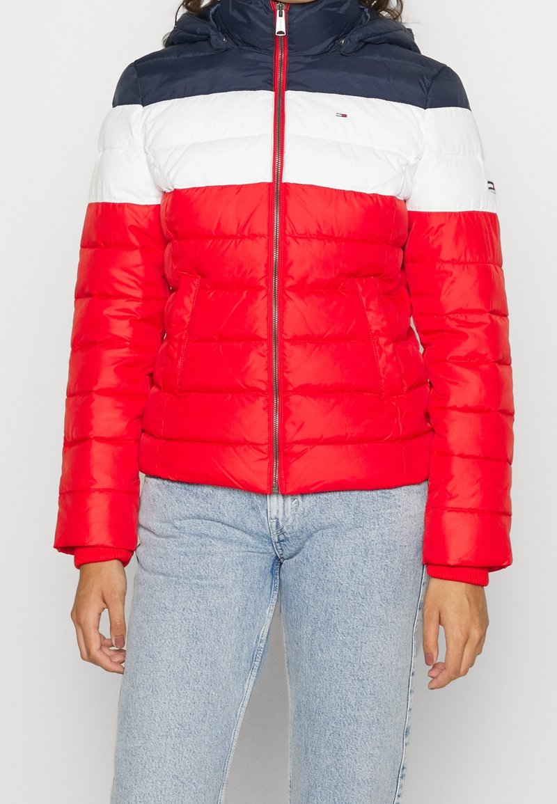 Person i en pufferjacka med marinblå, vit och röd horisontell randning och ljusblå jeans, stående mot en enfärgad bakgrund.