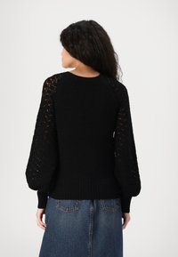 Maglione nero lavorato a maglia con maniche a palloncino in pizzo e orlo a coste; indossato con una gonna di jeans. Presenta un colletto tondo e dettagli testurizzati sulle maniche.