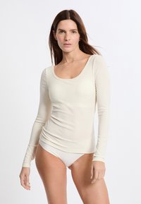 Donna con lunghi capelli castani che indossa un top aderente a maniche lunghe di colore crema e della biancheria intima abbinata su uno sfondo bianco.