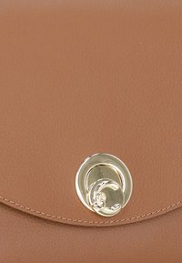 Sac à main en cuir marron avec une texture lisse, rabat arrondi et un détail de fermoir circulaire en or bien visible. Présente des coutures contrastantes.