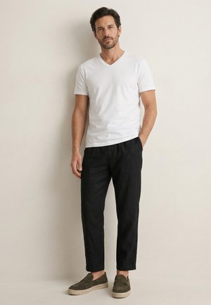 Homme debout contre un mur, portant un t-shirt blanc à col en V, un pantalon noir et des chaussures slip-on olive, la main dans la poche.