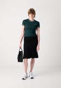 T-shirt ajustat de culoare verde închis cu margini albe, asortat cu o fustă neagră cu dungi. Modelul ține o geantă mică neagră și poartă adidași albi.