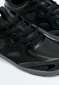 Gros plan de baskets athlétiques noires avec tissu en maille, superpositions texturées, lacets noirs épais et semelles en caoutchouc grises sur fond blanc.