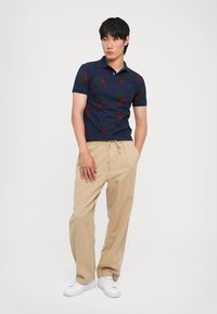 Ναυτικό πόλο μπλούζα με κόκκινες και πράσινες σχέσεις, κοντά μανίκια, σε συνδυασμό με άνετους μπεζ παντελόνες και λευκά αθλητικά παπούτσια.