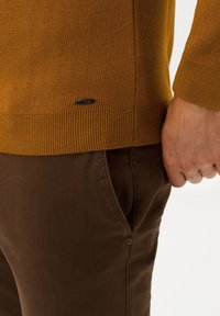 Nahaufnahme einer Person, die einen senfgelben Strickpullover und eine dunkelbraune Hose trägt, wobei ihre rechte Hand nahe der Tasche ruht.