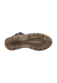 Semelle en caoutchouc brun avec un motif de grip texturé, présentant une légère courbure et une zone centrale pour le logo. Le matériau semble durable et flexible.