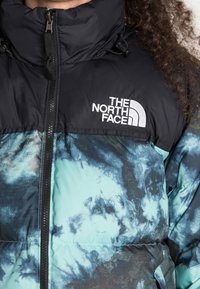 Gevoerde jas met een zwart bovenstuk en een blauw tie-dye patroon daaronder. Voorzien van een ritssluiting en een logo op de borst.