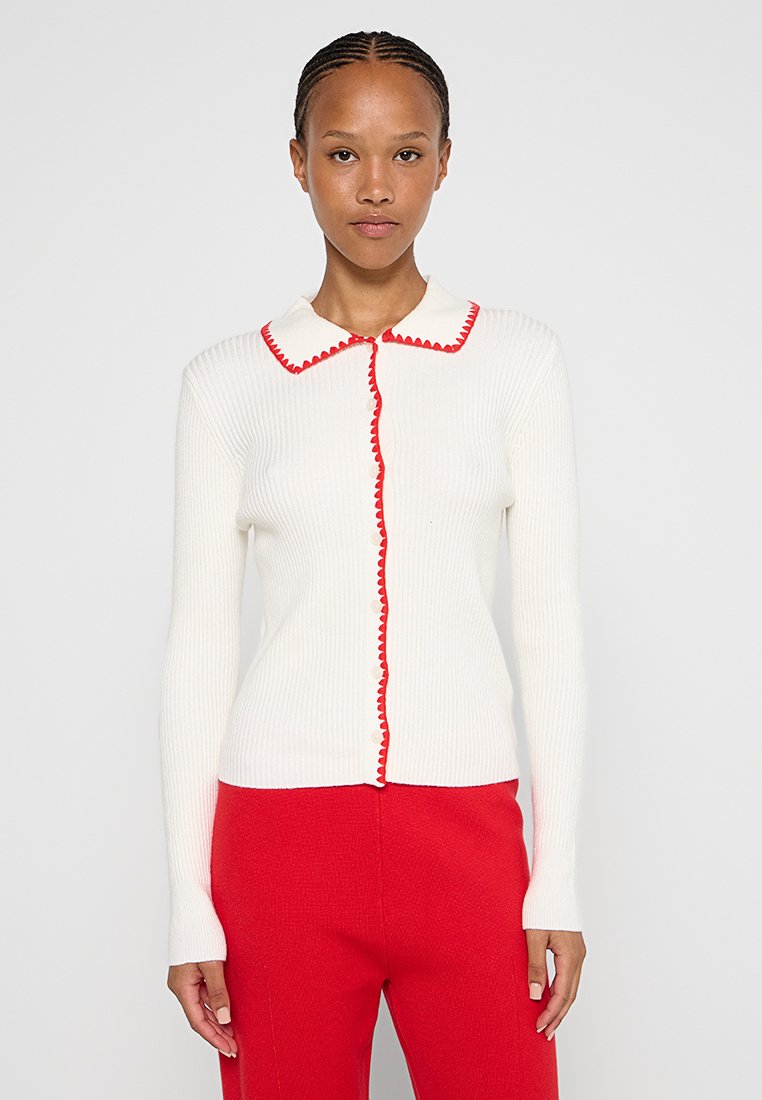 Weißer gerippter Pullover mit Kragen, rotem gewelltem Saum und durchgehendem Knopfverschluss. Getragen mit roten Hosen.