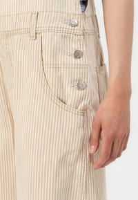 Salopette a righe beige con dettagli in metallo, tasche laterali e un tessuto testurizzato. Mano visibile appoggiata sul capo.