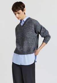 Grau gestrickter Pullover mit V-Ausschnitt, gerippten Bündchen, über einem hellblauen Hemd getragen, kombiniert mit dunklen Hosen. Glatte Textur, lässige Passform.
