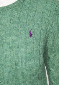Pull vert en maille torsadée avec un col rond. Présente un petit logo brodé violet, un motif en maille texturée et des poignets côtelés.