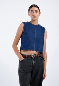 Top corto de mezclilla en azul oscuro, con mangas cortas y dobladillo sin acabar, presenta costuras en la parte frontal, combinado con jeans negros de tiro alto y cinturón marrón.