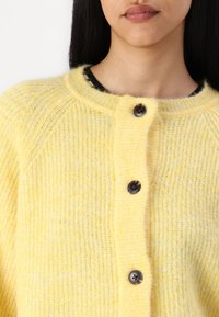 Cardigan en maille jaune avec un col rond, présentant une texture côtelée, trois boutons noirs et une finition légèrement duveteuse.