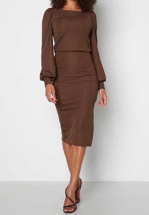 Femme portant une robe midi marron ajustée avec des manches longues bouffantes et des sandales à talons hauts noires à lanières, posant devant un fond blanc.