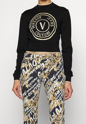 Sort langærmet kort sweater med stort beige "Versace Jeans Couture" logo, kombineret med sort, hvid og guldbelagt mønstret jeans med tekst og scroll-design.