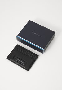 Tarjetero de cuero negro con diseño texturizado, cuatro ranuras y logotipo en relieve. Viene en una caja azul marina con una franja colorida.