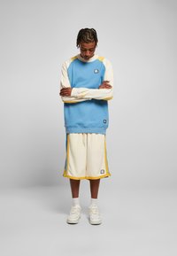 Lys blå hettegenser med gule og kremfargede detaljer, kombinert med kremfargede shorts med blå og gule sidelinjer og logo-patcher.