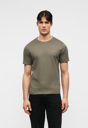 Vyras trumpais tamsiais plaukais dėvi alyvuogių žalios spalvos trumpomis rankovėmis marškinėlį be apykaklės ir juodus kelnes, stovintis prie šviesaus, paprasto fono.