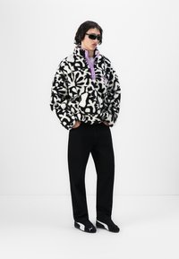 Zwart-wit gestreepte fleece trui met een paarse rits, gecombineerd met zwarte broek en zwarte schoenen, gedragen door een model met een zonnebril.