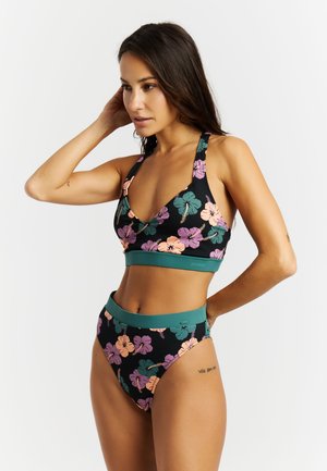 Vrouw die haar haar rechttrekt, draagt een zwarte, met bloemen bedrukte hoge taille bikini met groene rand, staand tegen een effen witte achtergrond.