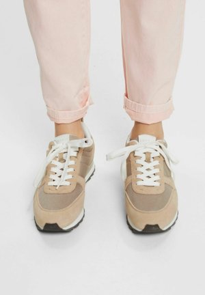 Pieds portant des baskets beiges et blanches à lacets assorties à un pantalon rose clair retroussé sur un fond blanc uni.